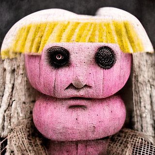 A pink, doll-like totem with hollow eyes, standing in a child’s place.