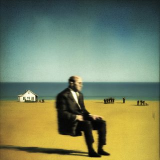 A motion-blurred man seated on a vast yellow shore, a distant house and small groups beyond.