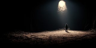 A single figure standing alone in a falling shaft of light in the dark.