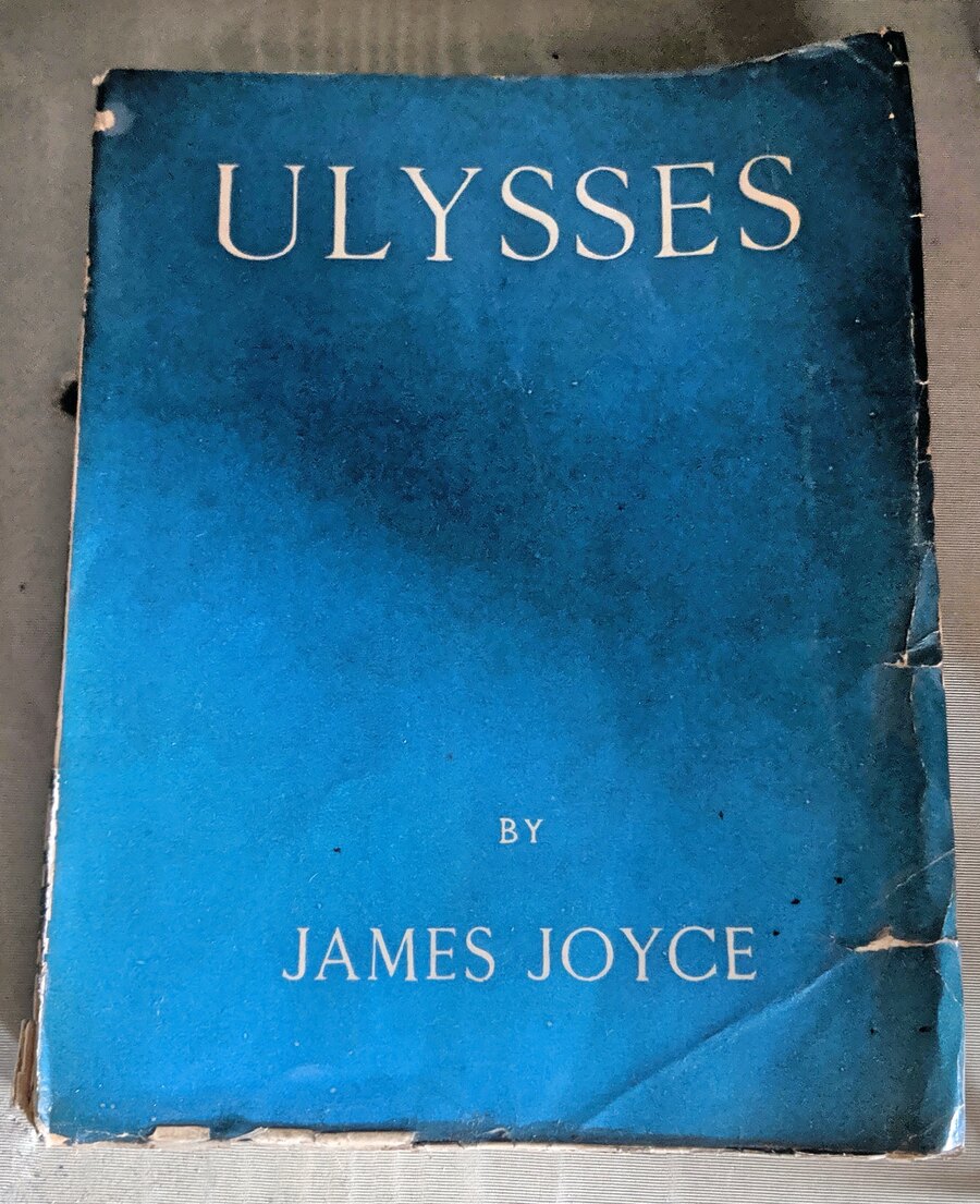 A worn blue-green paper-covered copy of the 1922 first edition of Ulysses stands upright in a museum display. White lettering reads Ulysses, James Joyce, Shakespeare and Company, Paris, 1922.