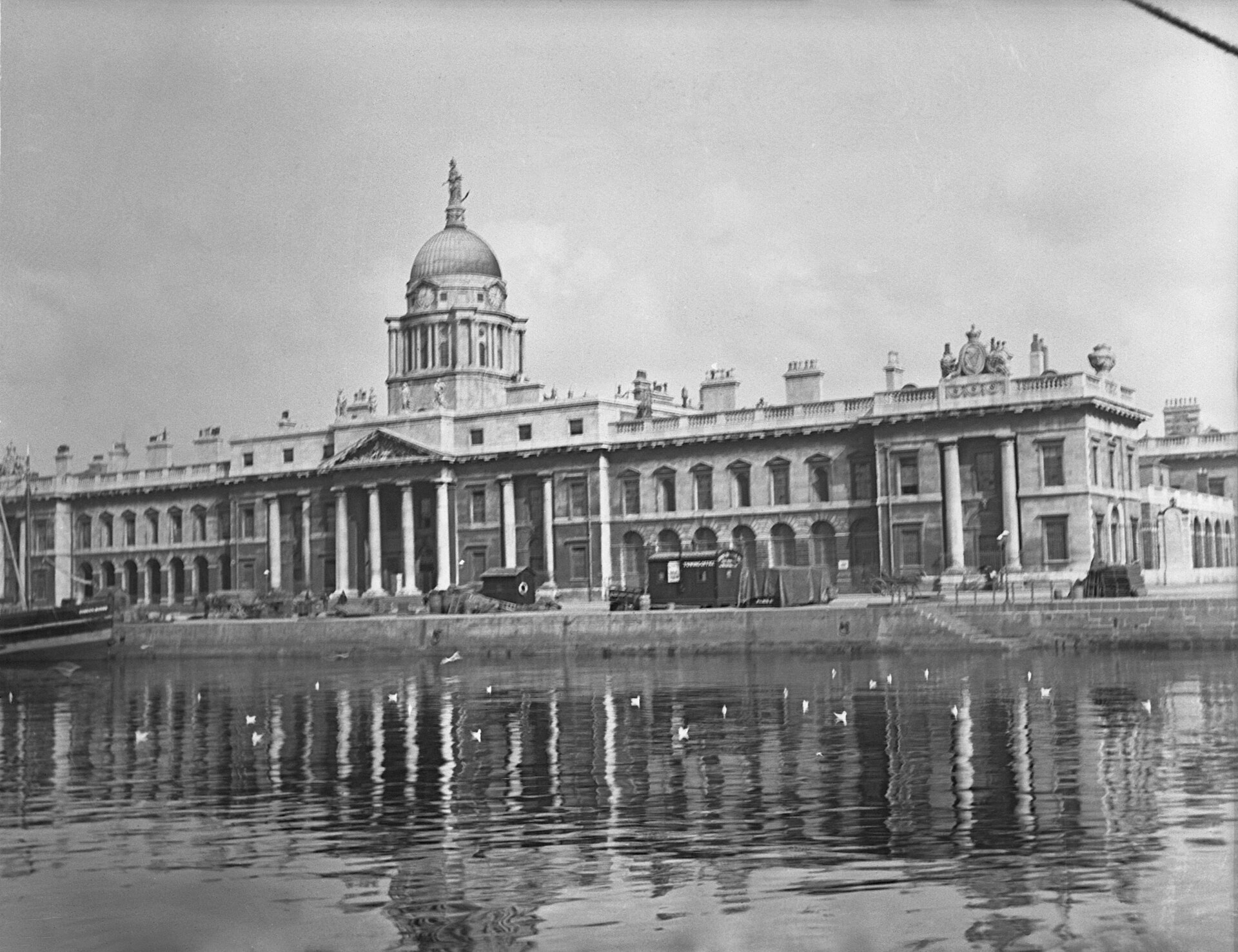 Black-and-white view across the River Liffey toward the Custom House, with quay walls and small boats in the foreground.