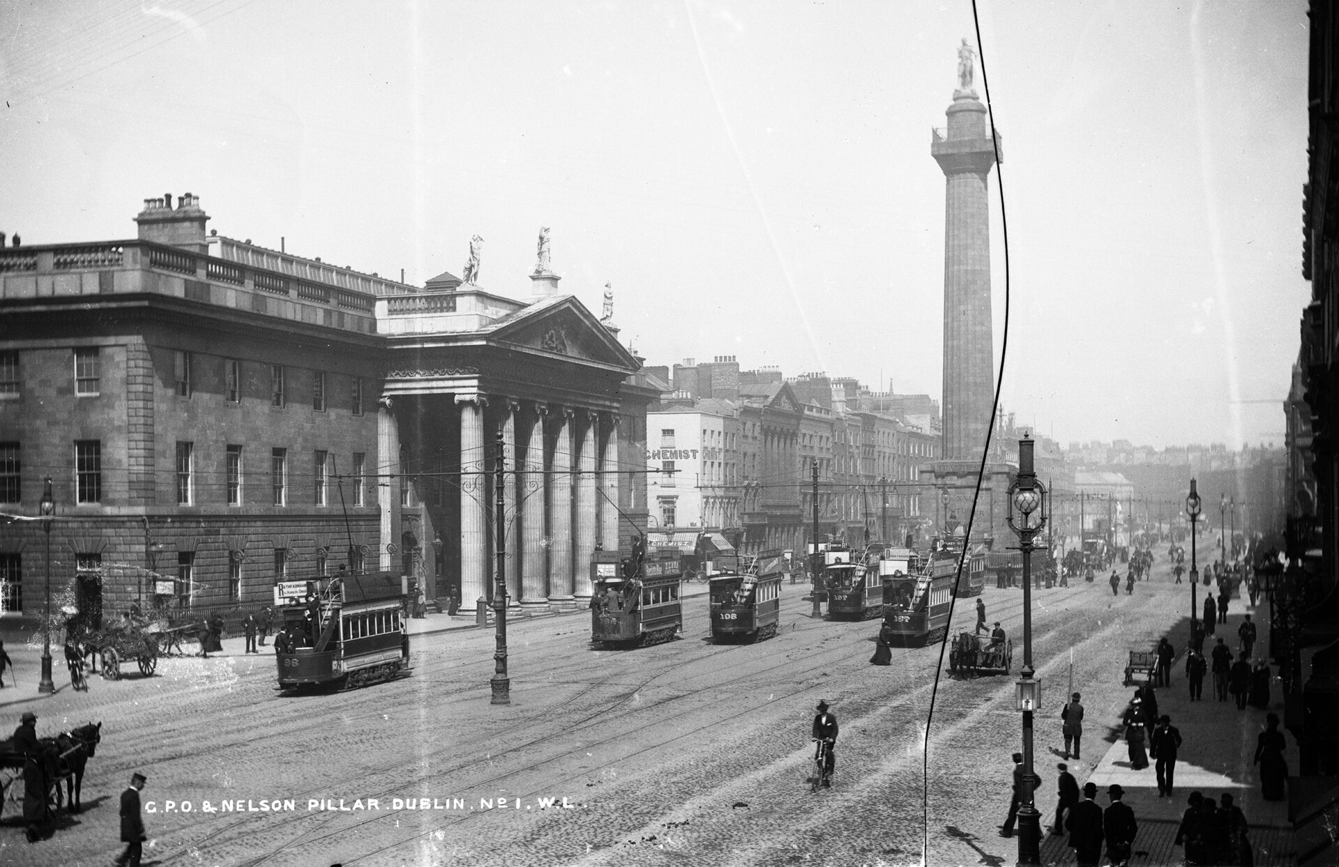 Black-and-white street view showing tram tracks, several trams, pedestrians, the General Post Office, and Nelson's Pillar on Sackville Street around 1900.