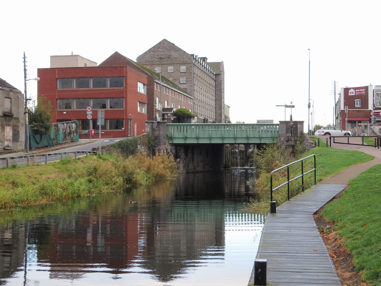 Still water of the Royal Canal fills the foreground; a narrow towpath curves beside it to a low green metal bridge, with grass and mixed industrial buildings beyond.