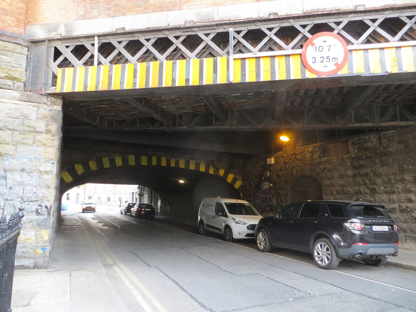 A narrow road passes beneath two stacked iron railway arches at Dublin Pearse station. Yellow-and-black height bars mark the lower arch; cars are parked under the stone and iron structure.