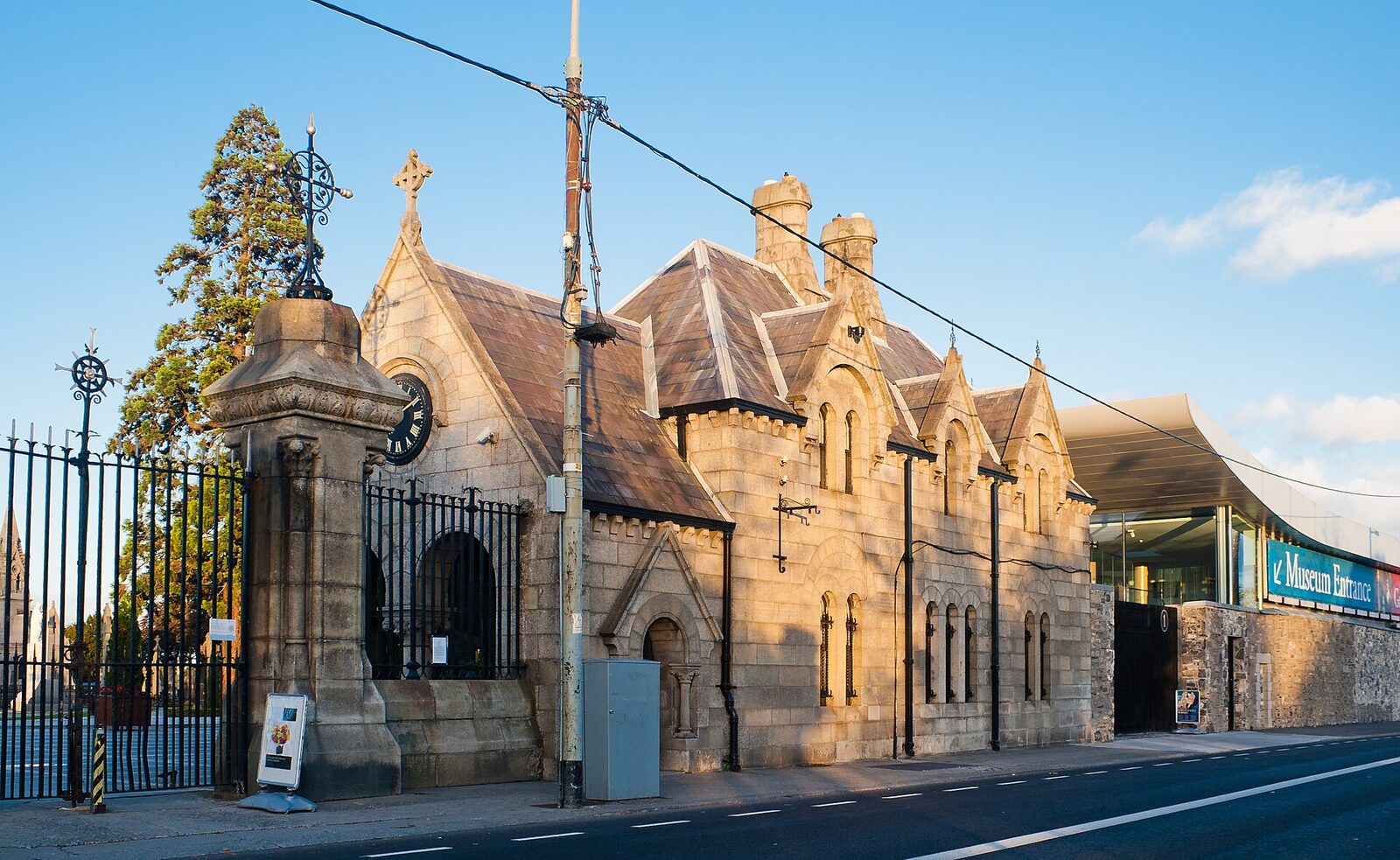 Stone Gothic cemetery entrance with clock and wrought-iron gates beside a modern glass museum entrance; the road and curb run across the foreground under a clear blue sky.