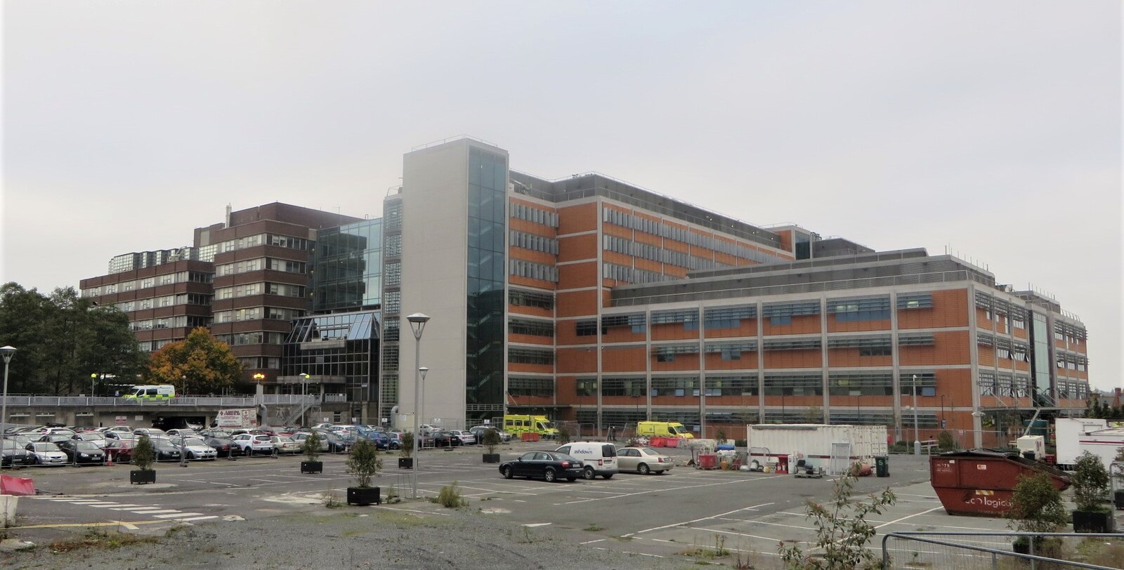 Wide view across a mostly open car park toward the Mater Hospital complex: low dark-brick blocks stand beside larger red-brick and glass buildings, with several yellow ambulances near the entrances under an overcast sky.