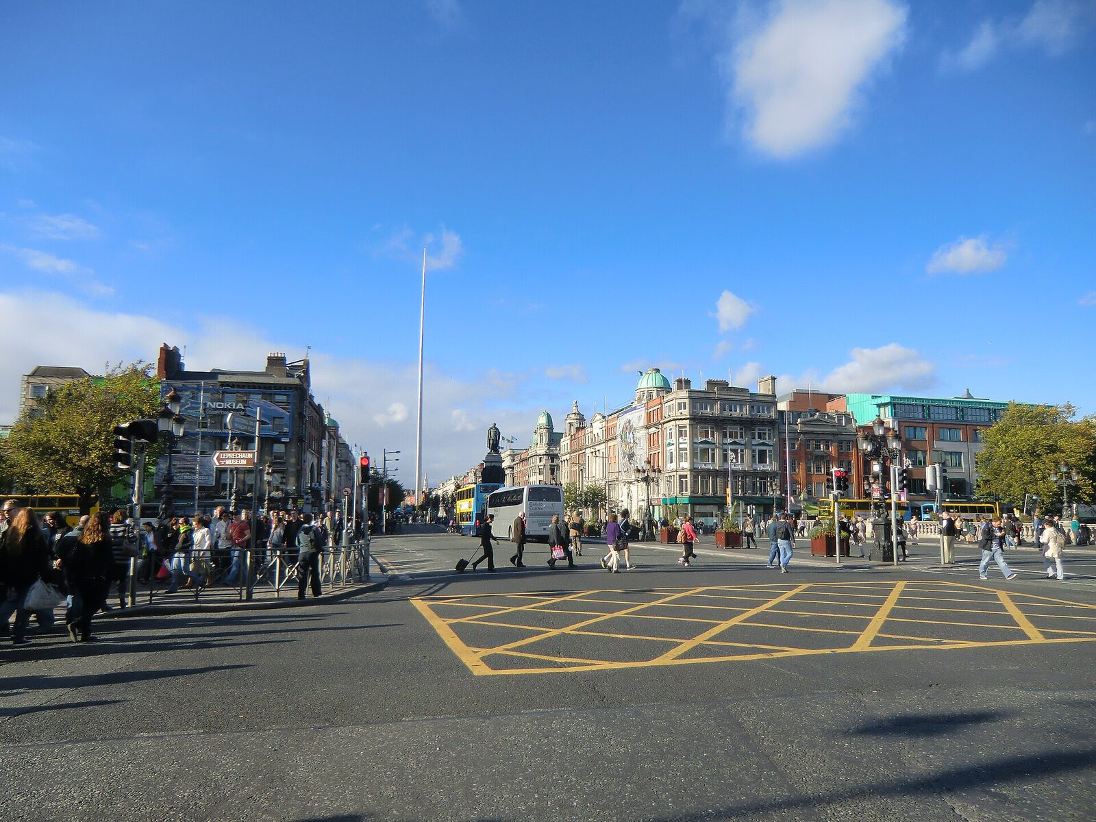 Daylight view north across the bridge’s wide roadway toward O’Connell Street. Pedestrians, buses, and traffic fill the junction; the O’Connell monument and Spire align down the centre.