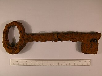 a hand-forged iron door key, dark, lying on a plain pale ground, its bow and wards in profile