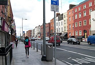 a wide view straight down a roadway, plain terraced houses on both sides, shopfronts at street level, parked cars along the kerb
