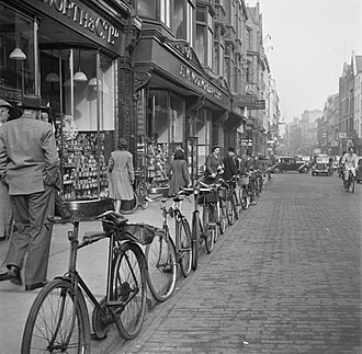 a rank of parked bicycles along a kerb in the foreground, a shopfront terrace running back on the left, figures on the pavement, traffic receding into haze