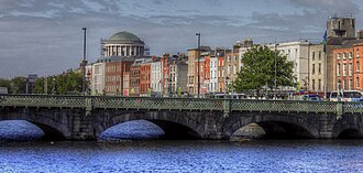 a river quay seen wide from across the water, a continuous terrace of tall houses along the waterfront, a green dome downstream